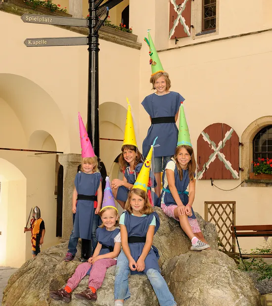 Kindergruppe in Kostümen mit bunten Hüten posiert auf Felsen an einem mittelalterlichen Ort